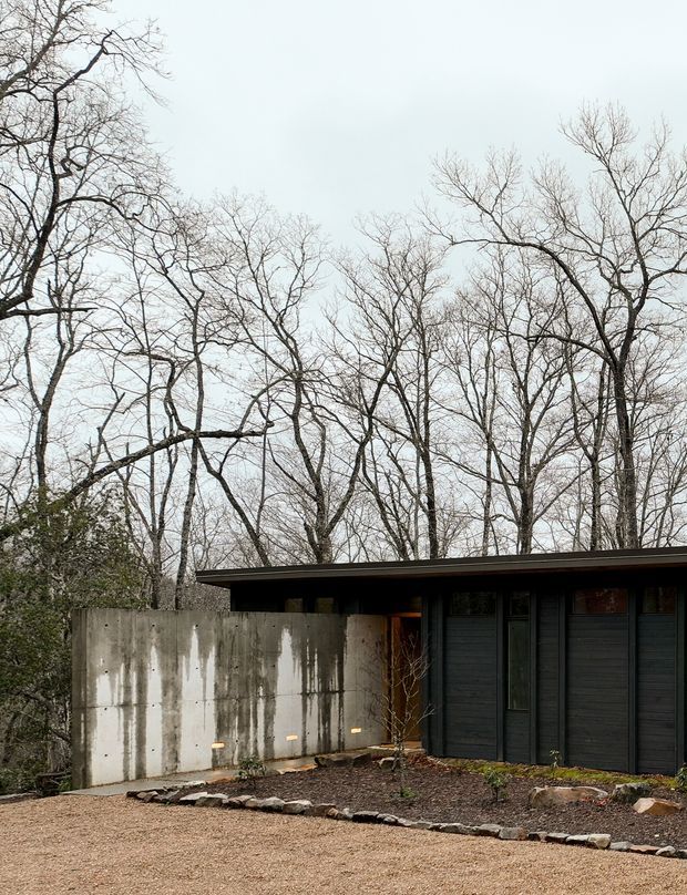 A low-profile modern house nestled among mature leafless trees. The dark horizontal facade has vertical cladding, narrow windows and a shallow projecting roof, while a weathered concrete privacy wall encloses a sparse gravel and stone-edged landscape. The overcast setting gives the exterior a quiet, restrained character.
