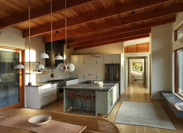 A warm, open-plan kitchen viewed from an adjoining dining area. The space features exposed timber ceiling beams and timber ceiling boards, white built-in cabinetry, a large pale island with integrated sink and seating, stainless-steel appliances, pendant lights, timber flooring, and generous glazed openings. A hallway leads toward a bedroom or living space in the background.