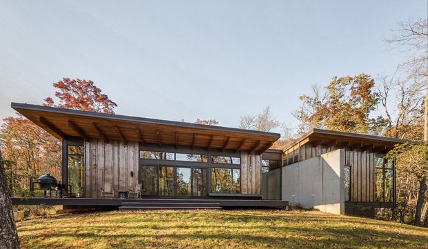 Exterior view of a low-slung contemporary house set among mature trees, composed of timber-clad volumes with broad projecting roof planes, extensive glazing, and a partially enclosed courtyard-like connection between wings. The warm timber soffits and autumn landscape create a natural, understated architectural character.