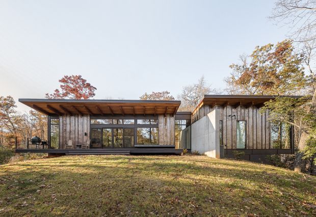 Single-storey modern residence set within a wooded suburban landscape, composed of two low rectangular volumes linked by a glazed central section. The exterior features vertical weathered timber cladding, broad projecting flat or very low-pitched roof planes with exposed timber soffits, extensive dark-framed glazing, and a simple concrete wall element. Autumn trees and a gently sloping lawn surround the house.