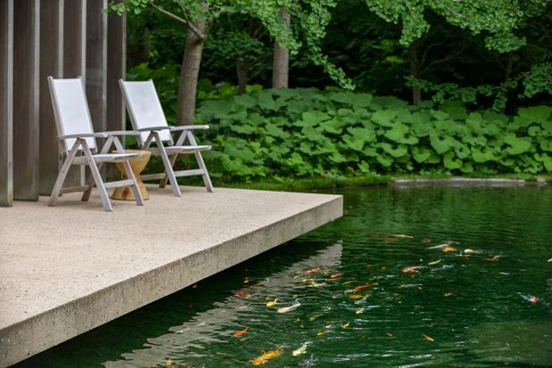 A minimalist concrete deck projects over a tranquil garden pond, beside a dark vertical-clad structure. Two white reclining outdoor chairs sit on the deck, with dense leafy vegetation and colourful fish visible in the water. The scene has a calm, nature-integrated outdoor retreat character.