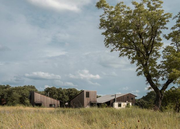 A low-profile rural architectural complex viewed across a grassy field, composed of several connected timber-clad buildings with pitched and shed roofs. Mature trees, native vegetation, and an overcast sky frame the scene, giving the property a restrained, agrarian character.