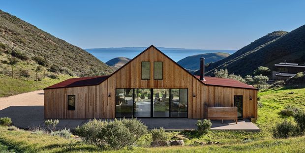 A compact single-storey timber-clad house set in a steep mountain valley. The symmetrical gabled central volume has large glazed doors or windows, flanking low wings with red standing-seam metal roofs, and a small outdoor bench. The surrounding landscape features native vegetation, exposed gravel, and dramatic hills beneath a clear blue sky.