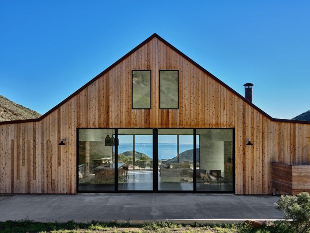 Front exterior view of a contemporary gable-roofed building clad in vertical natural timber boards. A large black-framed glazed opening reveals a light-filled interior, while two upper windows and a roof chimney punctuate the symmetrical facade. A concrete terrace sits in front, with rugged hills visible through the glazing and around the building.