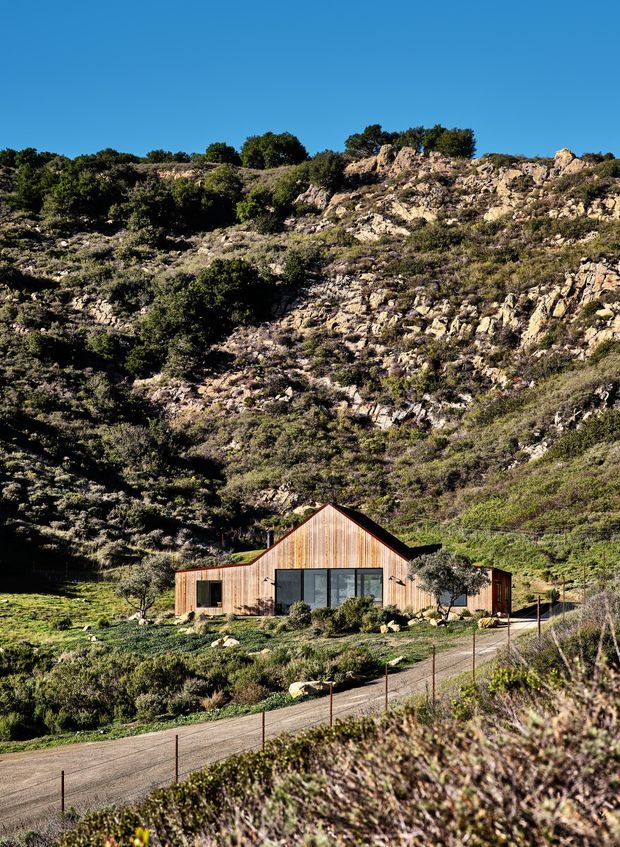 A compact single-storey timber-clad house set against a rugged, rocky hillside in a dry rural landscape. The home has a simple gabled central volume, low side wings, dark-framed glazing, and a modest gravel access road and fenced foreground. The image is an exterior architectural photograph under clear blue sky, with the surrounding bush-covered rock escarpment dominating the setting.