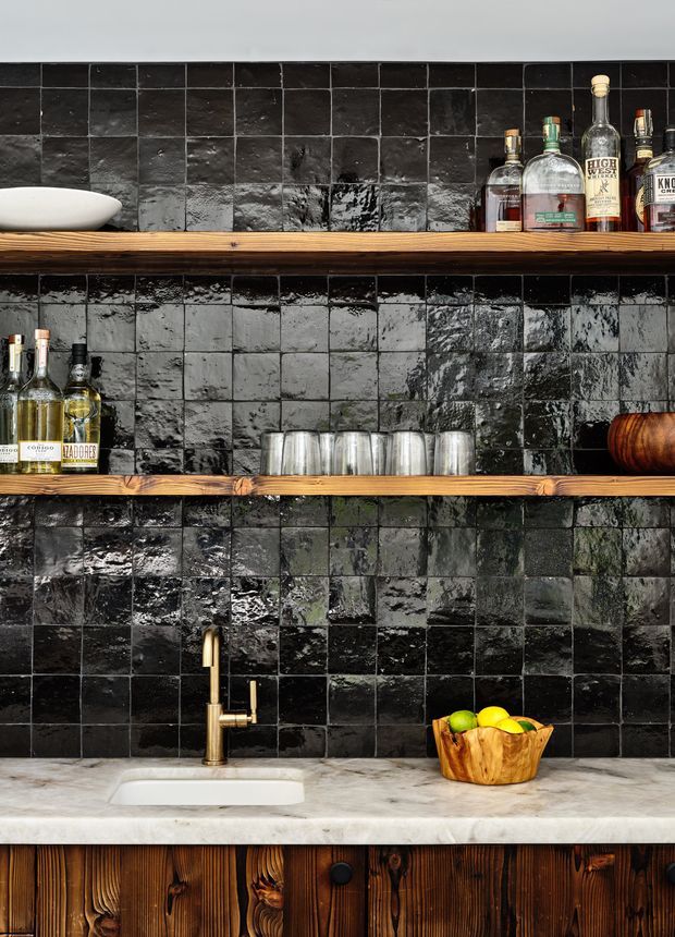 Close-up view of a compact bar or kitchenette wet area with glossy, irregular black square tile splashback, thick rustic timber open shelves holding liquor bottles and glassware, a white vessel sink with brass gooseneck tap, pale stone benchtop, and dark stained timber cabinetry below. The composition is richly textured and moody, with warm wood and brass contrasting against the black tile.
