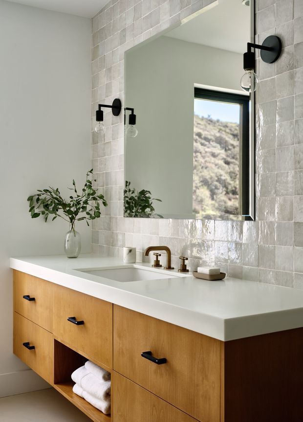 A bright, refined bathroom featuring a wall-mounted timber vanity with black pulls, a white solid-surface benchtop, integrated basin, and brushed brass tapware. The backsplash and surrounding wall are clad in small glossy pale grey-beige tiles, with a large black-framed mirror and matching black wall sconces. A simple vase with greenery and neatly folded towels add softness.