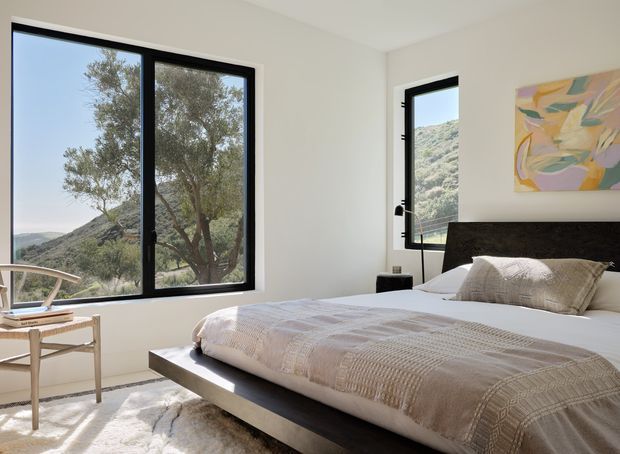 Bright, contemporary bedroom with a large platform bed, dark upholstered headboard, layered neutral bedding, and a small light-wood desk and chair beside expansive black-framed windows. The windows overlook a dry, hilly landscape with mature trees. A colourful abstract artwork adds a restrained accent above the bed.