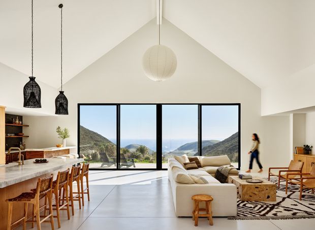 Open-plan contemporary living space with a kitchen and dining area on the left and a living room on the right. The room has a dramatic vaulted ceiling, large black-framed floor-to-ceiling glazing, pale walls, polished light flooring, a stone kitchen island, timber stools and chairs, pendant lighting, and a large cream sectional sofa. Mountain and water views are visible beyond the glazing. Natural timber furnishings and a patterned rug add warmth to the bright interior.