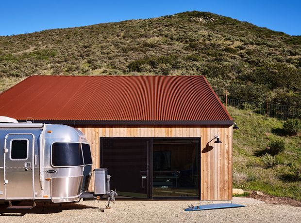 Exterior view of a compact timber-clad structure with a rust-red corrugated metal pitched roof, large dark-framed glazed sliding door, and exterior wall light. A silver travel trailer is parked in front, with a scrub-covered hillside rising directly behind the building.