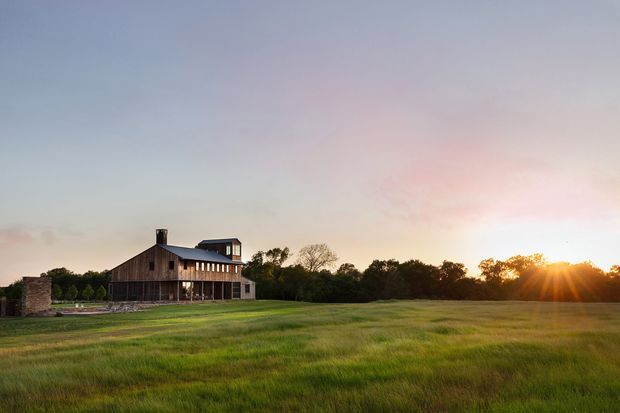 A wide exterior view of a substantial two-storey rural residence set within an expansive grassy field at sunset. The building has dark timber weatherboard cladding, a long low roofline, a small rooftop structure, a stone chimney or wall element, and extensive horizontal glazing along the upper level. Mature trees form a backdrop, while warm evening sunlight creates a serene pastoral atmosphere.