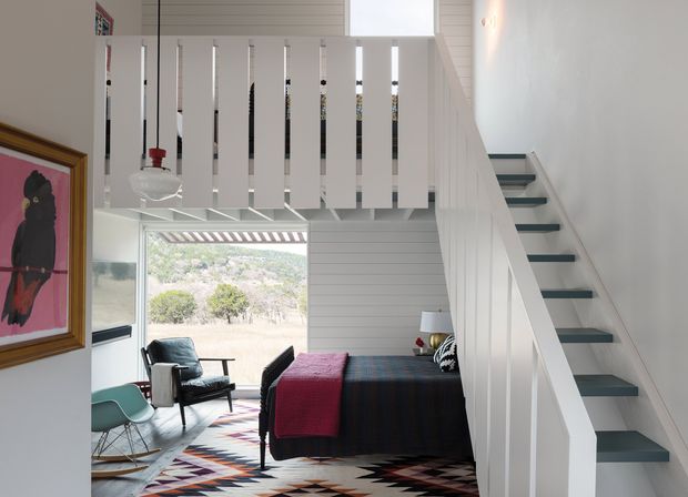 Bright, compact open-plan interior showing a staircase rising beside a bedroom or sleeping area. The stair has white painted vertical balustrades and dark treads, while the lower level includes a bed, lounge chairs, a patterned rug, framed artwork, and a pendant light. Large glazing opens the space to a leafy landscape, and white horizontal wall cladding adds texture.