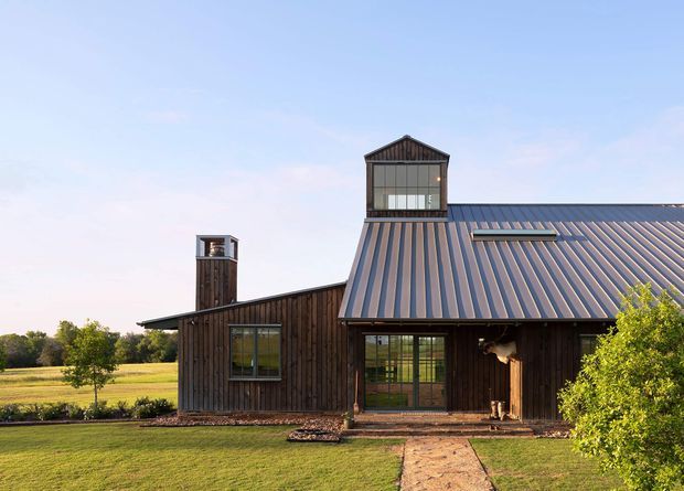 Exterior view of a single-storey barn-like home set in an open rural landscape. The building has dark vertical timber cladding, a large pitched standing-seam metal roof, glazed dormer-like clerestory structures, broad timber-framed windows and doors, and a simple path leading to the entrance. The composition has a warm, rustic yet carefully designed character.