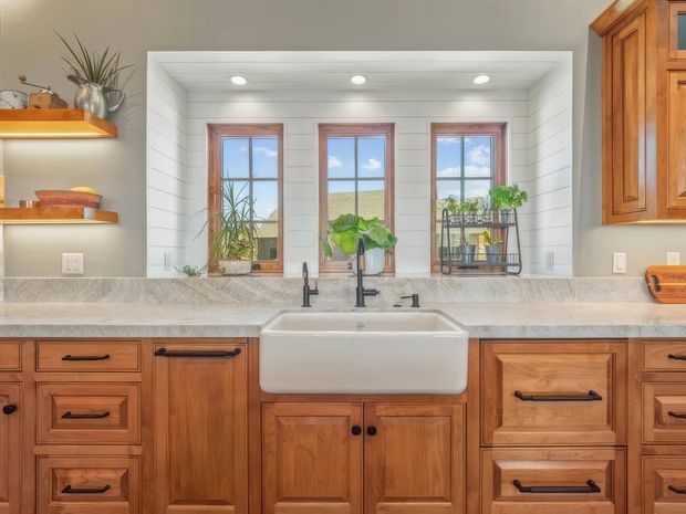 Bright, traditional-style kitchen featuring warm wood cabinetry, a large white farmhouse apron-front sink, marble-look benchtops, matte black tapware and hardware, and open shelving. A recessed bank of three windows is framed with white horizontal wall panelling, bringing in views of the landscape and sky. Plants and simple accessories add a natural touch.