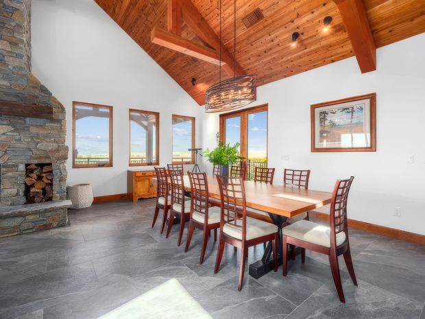 Bright, spacious dining room with a long wooden dining table and numerous upholstered wooden chairs. The room features a dramatic vaulted timber-lined ceiling with exposed structural beams, white walls, slate-like grey flooring, and a large natural-stone fireplace at the left. Multiple timber-framed windows provide expansive mountain and landscape views. A sculptural woven pendant light hangs above the table, while a timber sideboard and potted plant add warmth.