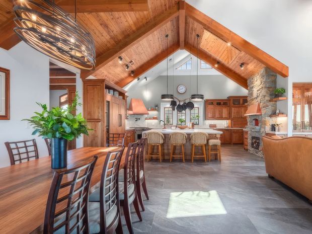 Spacious open-plan great room with a dining area in the foreground, a large kitchen with island seating beyond, and a living area to the right. The room features a dramatic exposed timber cathedral ceiling with heavy beams, warm timber cabinetry and furniture, grey stone-look flooring, a substantial stone fireplace, and abundant natural light. A sculptural metal pendant hangs over the dining table, while additional pendant lights illuminate the kitchen island.