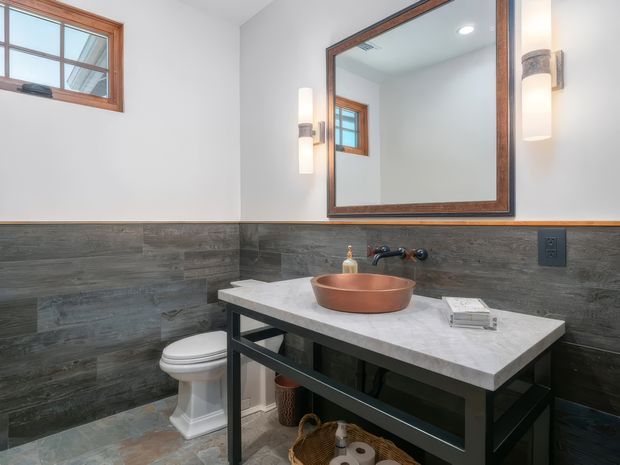 A bright, compact bathroom with a floating-style dark-framed vanity, pale stone benchtop, copper vessel basin, wall-mounted dark tapware, and a white toilet. Dark grey wood-look wall tiles cover the lower walls, complemented by a mottled tiled floor and white upper walls. A large timber-framed mirror, matching timber-framed window, and contemporary wall sconces add warmth and contrast.