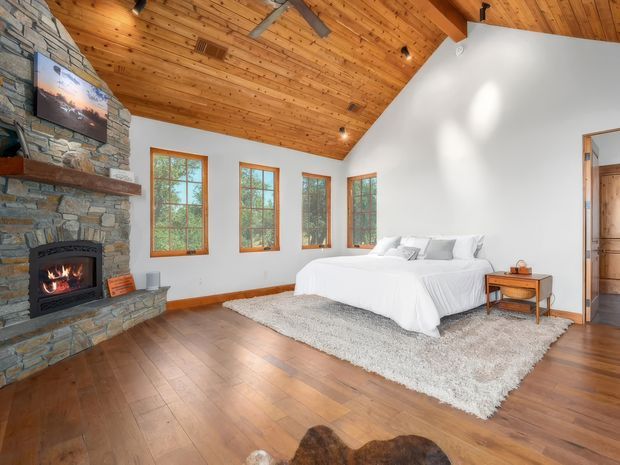 A spacious rustic bedroom with a white bed positioned beneath a high sloped timber ceiling. The room features warm timber flooring, exposed timber ceiling boards and beams, natural stone wall cladding, and a substantial stone fireplace with a lit fire. Multiple timber-framed windows provide views toward trees, while a light area rug, small bedside table, and simple bedding create a warm, relaxed retreat.