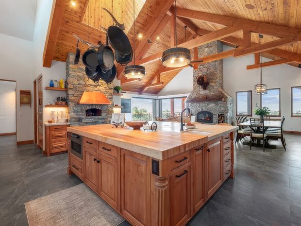 Spacious rustic kitchen and dining area dominated by a large timber island with a pale stone benchtop, integrated appliances, farmhouse-style cabinetry, and a sink. The room features a vaulted timber ceiling with exposed heavy beams, multiple pendant lights, a stone-clad fireplace and chimney, hanging cookware, and large windows. A dining table is visible beyond the kitchen.
