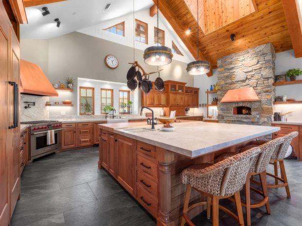 Spacious, highly detailed kitchen with a large central island, extensive warm timber cabinetry, woven bar stools, stainless-steel appliances, and a substantial natural-stone fireplace surround. The room features a vaulted timber-lined ceiling, clerestory-style windows, pendant lighting, dark tile flooring, and a warm rustic lodge-inspired character.