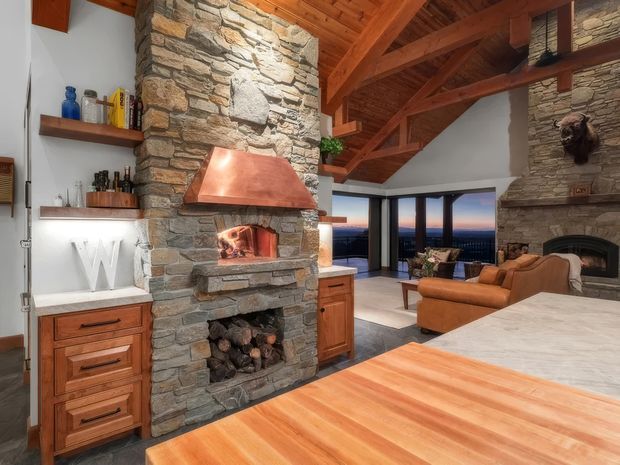 A rustic open-plan kitchen and living area centered on a substantial natural-stone fireplace and copper range hood. The kitchen includes warm timber cabinetry, open shelving, and a large timber island with a pale stone benchtop. Exposed timber beams and a timber-lined vaulted ceiling extend through the living space, which features leather seating, a second stone fireplace, and broad glazing with a distant view.