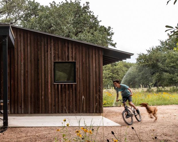 A compact single-storey timber-clad outbuilding with a low mono-pitched roof and small dark-framed window, set in a landscaped rural garden. A cyclist and dog pass in the foreground, while mature trees and flowering meadow planting surround the structure.