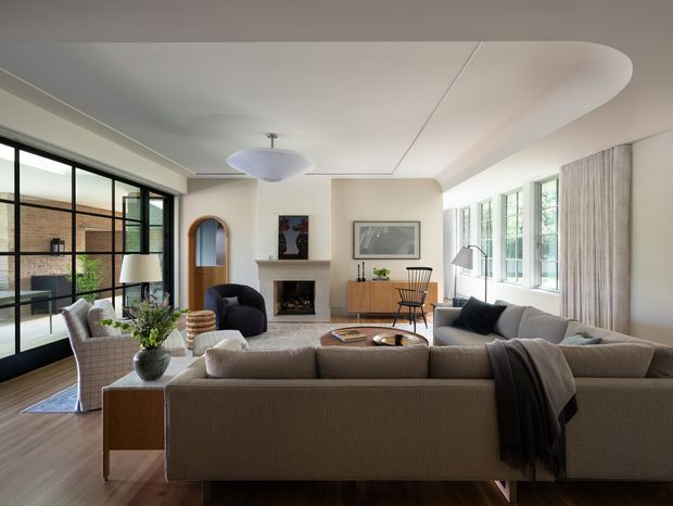 A bright, refined living room with a large sectional sofa arranged around a central coffee table and fireplace. The space features dark timber flooring, pale upholstered furniture, a sculptural pendant light, extensive black-framed glazing, a curved ceiling detail, built-in cabinetry, and an arched timber doorway. A courtyard or adjacent outdoor area is visible through the glazing.