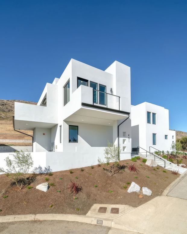 A contemporary white stucco multi-level residential house with crisp geometric volumes, projecting balconies, large dark-framed windows, and a flat roofline. The home sits on a dry, landscaped sloping site with sparse drought-tolerant planting, rocks, concrete paths, and a suburban street frontage under a clear blue sky.