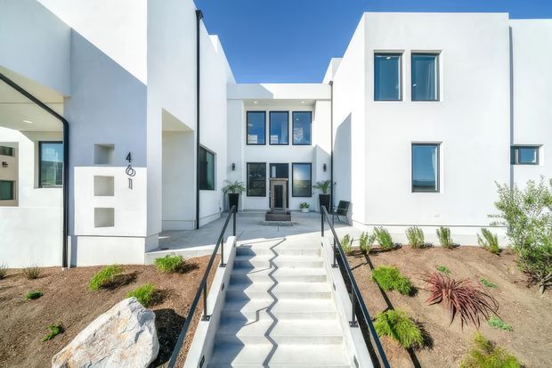 Exterior view of a bright white modern multi-storey residential building arranged around a central entry courtyard. The approach features broad concrete steps with a dark zigzag inlay or shadow pattern, black metal handrails, sparse contemporary landscaping, large rocks, and recessed glazing. Strong geometric forms, flat rooflines, and blue-sky contrast create a clean, minimalist architectural composition.