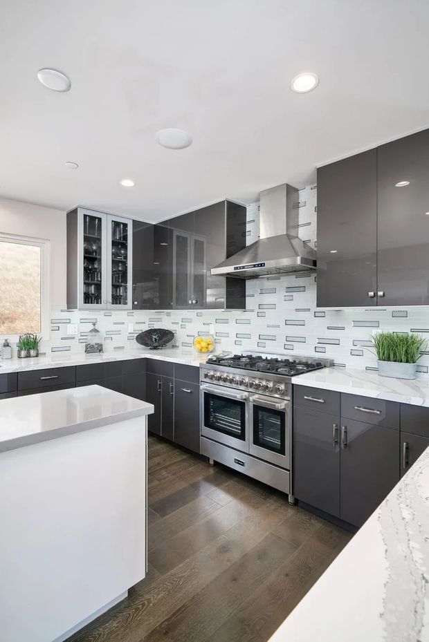 Bright contemporary kitchen with dark charcoal-grey flat-panel cabinetry, extensive white benchtops, a large central island, stainless-steel range cooker and hood, and a white rectangular-pattern tiled splashback. Warm timber flooring contrasts with the cool cabinetry, while glass-front display cabinets and recessed ceiling lights complete the fitted kitchen.