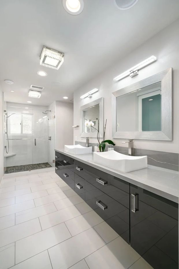Bright contemporary bathroom featuring a long double vanity with white vessel sinks, dark grey flat-panel cabinetry, a light grey stone benchtop, large framed mirrors, white tiled walls and floor, and a glass-enclosed shower at the rear. Recessed and linear vanity lighting create a clean, well-lit interior.