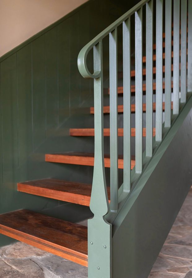 Close-up architectural photograph of an interior staircase with dark timber treads, painted deep green wall panelling and a matching green metal balustrade. The balustrade features slender vertical spindles and a distinctive curved, ornamental handrail end. A slate- or stone-look floor is visible at the base, creating a refined, moody traditional character.