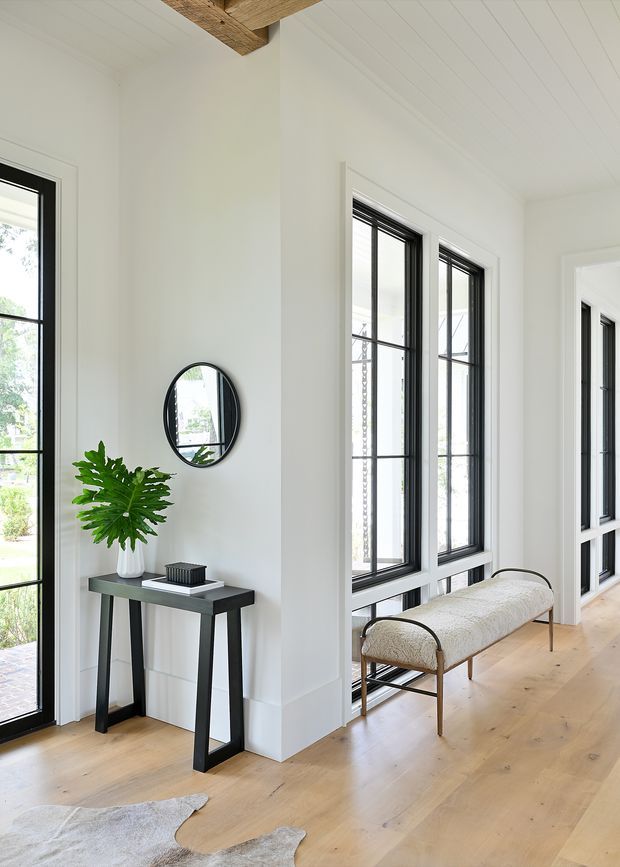 Bright, refined residential hallway or entry space with white paneled walls and ceiling, tall black-framed windows and glazed doors, wide light timber flooring, and exposed timber ceiling beams. A slim dark console table with a potted plant and round mirror sits beside the glazing, while a cushioned bench occupies the window-lined corridor. The space has abundant natural light and a clean contemporary-traditional character.