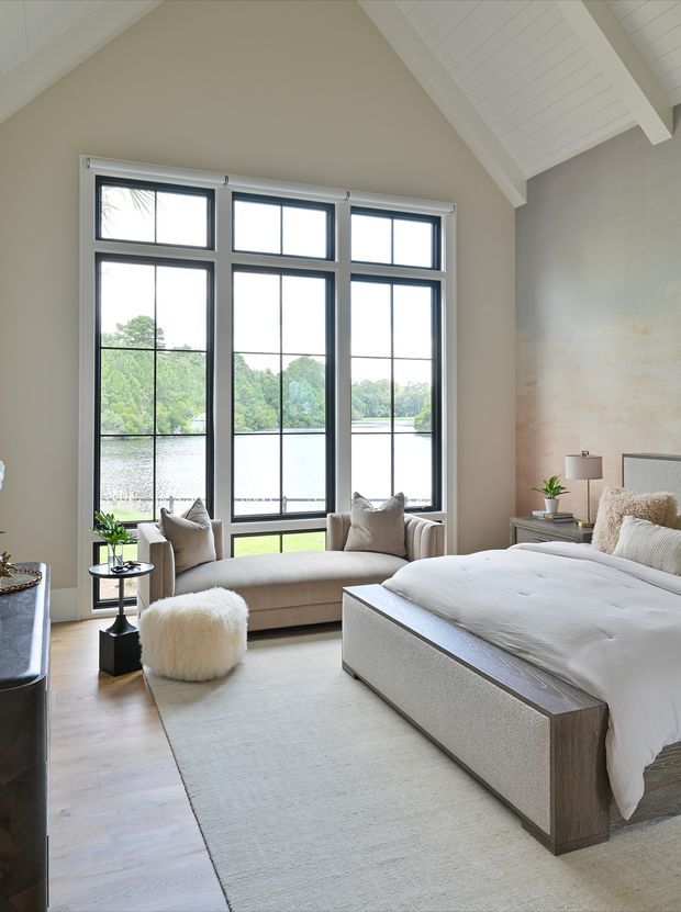 A bright, refined bedroom with a large upholstered bed, neutral bedding, a soft area rug, and a compact seating area positioned in front of expansive black-framed windows. The room has a vaulted ceiling with white painted timber paneling and exposed beams, pale walls, light timber flooring, and views across a body of water and greenery. Furnishings include a bench-style sofa, accent chairs, a small side table, bedside tables, lamps, and a textured ottoman.