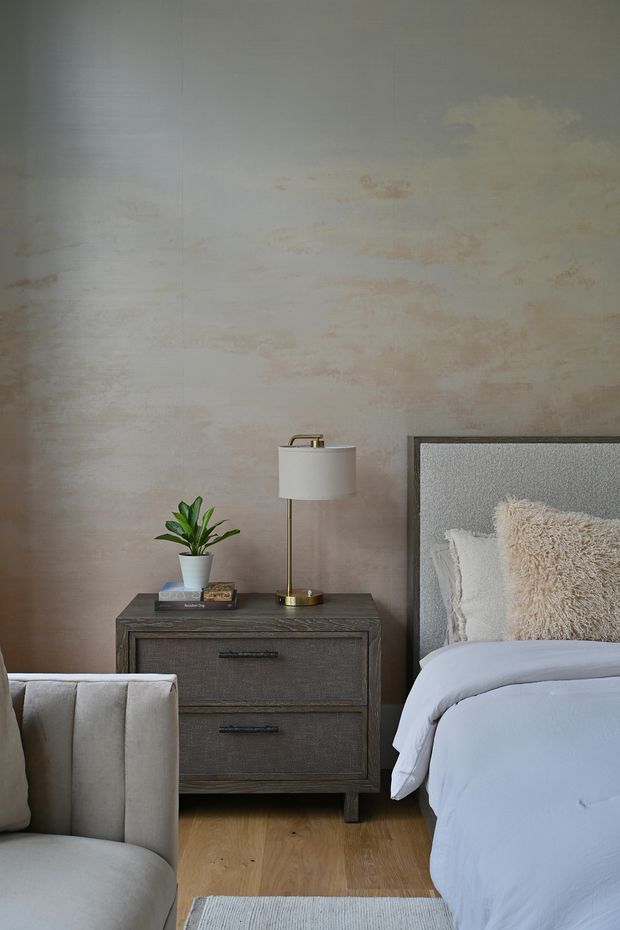 A softly styled bedroom corner featuring a partially visible upholstered bed with pale blue-grey bedding, a textured grey upholstered headboard, and a dark wood bedside table with two drawers. A brass table lamp, books, and a small potted plant sit on the nightstand. The room has warm light timber flooring, a mottled beige plaster-like wall finish, and a neutral upholstered chair in the foreground.
