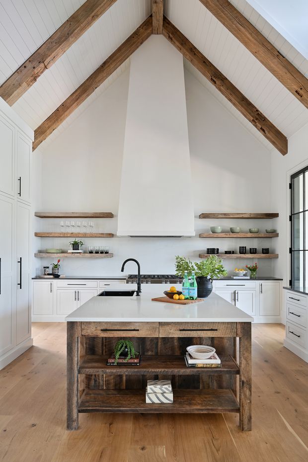 Bright, spacious kitchen with a dramatic vaulted white-panelled ceiling and exposed rustic timber rafters. White Shaker-style cabinetry lines the walls, with open reclaimed-wood shelves, a large white central island benchtop, dark stone-looking perimeter benchtops, and a substantial rustic timber island base with open shelving. Tall black-framed glazing is visible on the right, while warm timber flooring and timber accents add contrast.
