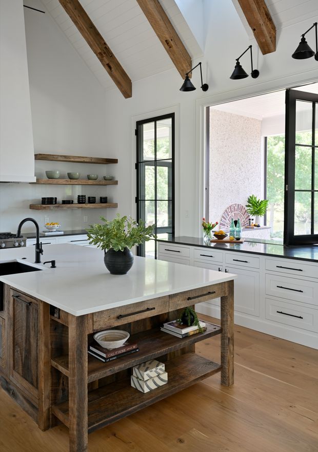 Bright, spacious kitchen with a large white island featuring a rustic timber base and open shelving. White shaker-style cabinetry lines the rear wall beneath a dark countertop, while floating reclaimed-wood shelves display ceramics and glassware. Black-framed windows and glazed doors provide abundant natural light. The ceiling has white boards with exposed rustic timber beams, complemented by black pendant and wall-mounted lights. The overall design blends modern farmhouse and contemporary elements.