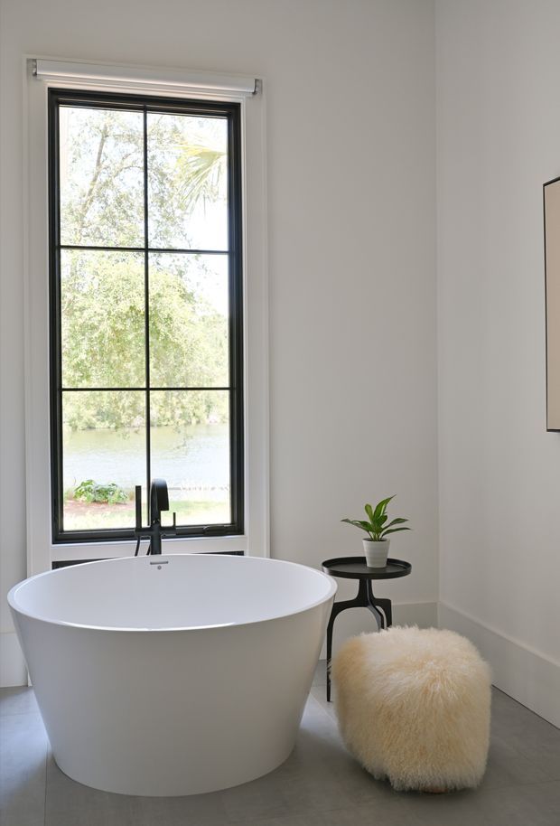 A bright, refined bathroom featuring a freestanding white oval bathtub positioned beside a tall black-framed multi-pane window. A matte-black wall-mounted bath filler rises behind the tub, with a small black round side table holding a potted plant and a soft cream boucle stool nearby. Light neutral walls and grey flooring create a calm, minimalist setting, with leafy waterfront scenery visible through the window.