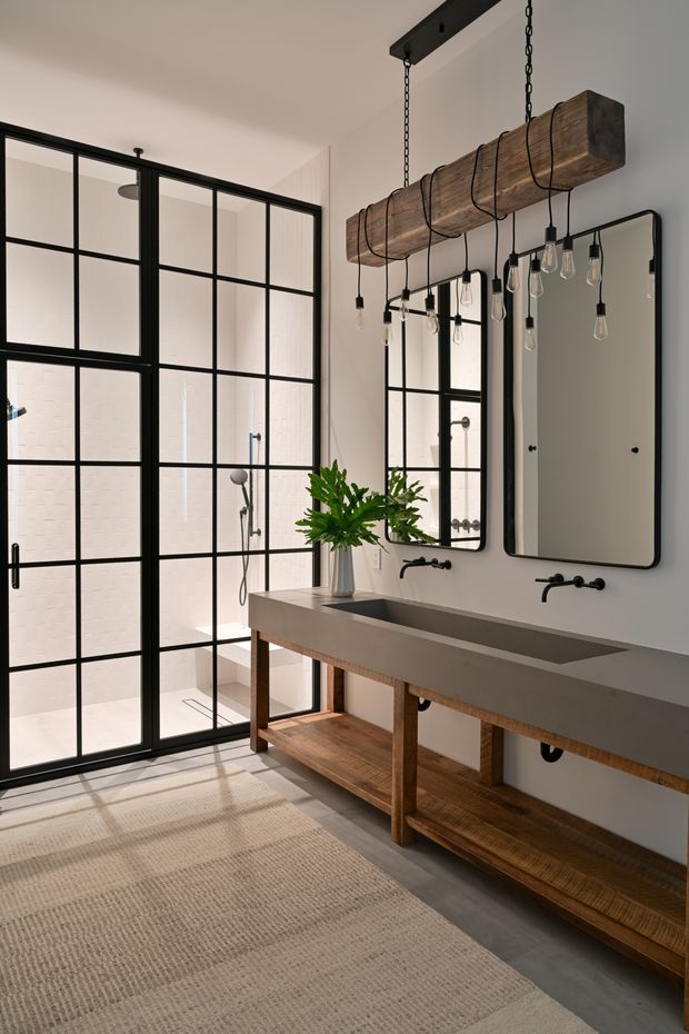 A spacious, refined bathroom featuring a long double vanity with a grey stone-like benchtop, open timber shelving, two wall-mounted black faucets, and two rounded-rectangle mirrors. A large black-framed steel-look glazed shower enclosure dominates the left side. Above the vanity is a dramatic suspended timber beam light fitting with exposed pendant bulbs and black chains. The room has warm white walls, dark grey flooring, natural timber, black metal details, and a neutral woven rug, creating an industrial-modern, warm aesthetic.