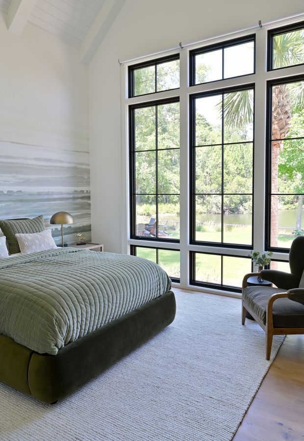 A serene, light-filled bedroom with a large upholstered bed in muted sage green, layered bedding, and a textured pale rug over timber flooring. Floor-to-ceiling black-framed grid windows provide expansive views of trees, lawn, and a pond. The room includes a sculptural bedside lamp, a textured feature wall, and a timber-framed lounge chair. The ceiling has a white pitched, timber-lined form with exposed beams.