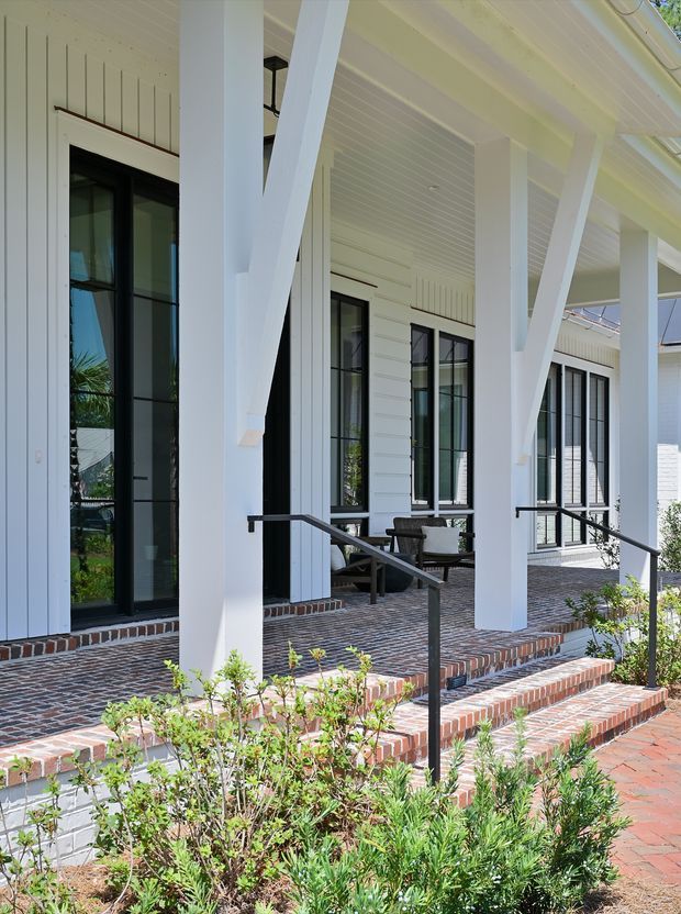 A bright covered front porch with prominent white timber posts and diagonal braces, white vertical and horizontal wall cladding, black-framed glazed doors and windows, and a beadboard-style ceiling. Brick paving forms the porch floor, steps, and low edging walls, with simple black metal handrails and landscaped shrubs in the foreground. The image presents a refined traditional coastal-style residential exterior and entry approach.