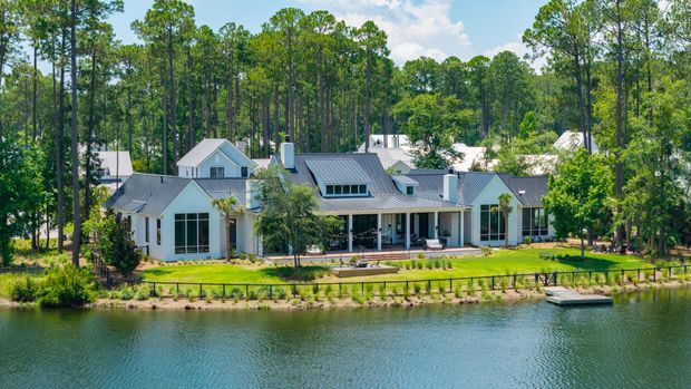 A large, low-rise residential home viewed across a body of water, set within a heavily wooded site. The house has white exterior walls, multiple pitched gabled roof forms with dark metal roofing, extensive glazing, covered outdoor verandas, and landscaped lawn leading to a small waterside dock. Tall trees surround the property, creating a secluded waterfront setting.