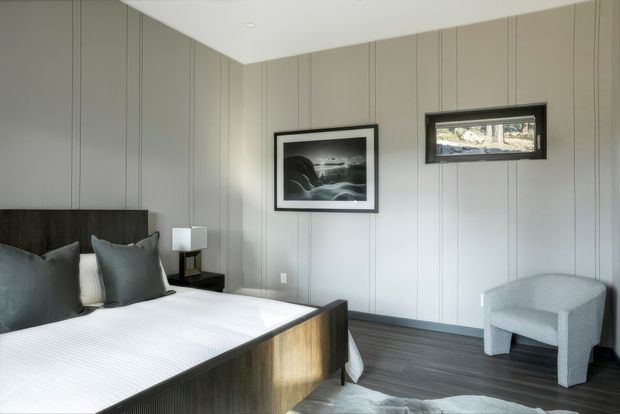 A contemporary bedroom with a low-profile timber bed featuring a dark wood headboard and footboard, grey bedding and pillows, vertically panelled grey walls, dark timber flooring, a small bedside table and lamp, framed monochrome artwork, and a light grey upholstered accent chair. The room has a clean, restrained, neutral palette and recessed ceiling lighting.