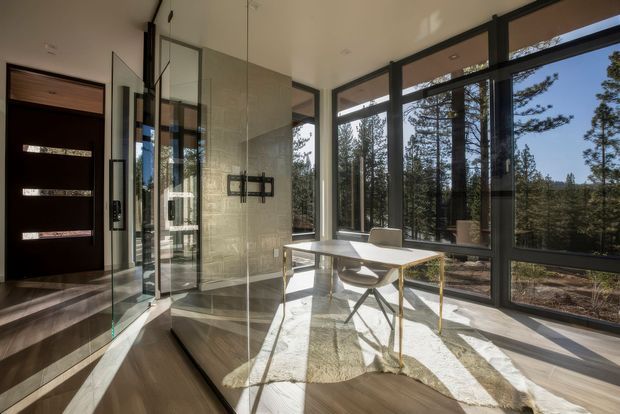 Bright, minimalist contemporary interior with a compact dining or work table beside floor-to-ceiling black-framed glazing overlooking tall trees. A glass-partitioned entry or hallway, dark timber front door, pale tiled wall, light timber flooring, and sculptural white chairs create a clean, refined space with strong natural light and long shadows.