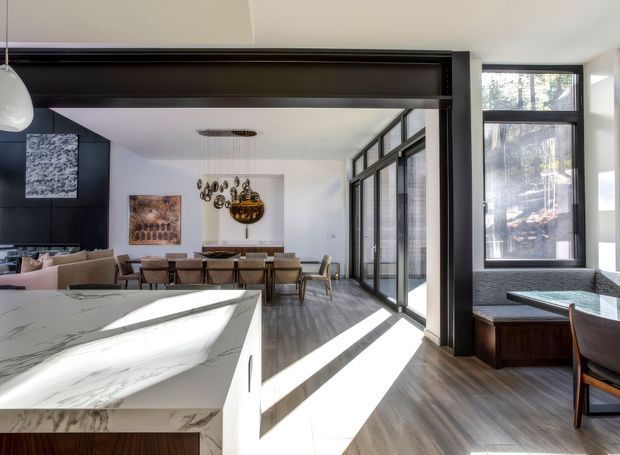 Bright open-plan contemporary interior showing a kitchen island in the foreground, a dining area beyond, and a living area to the left. Full-height black-framed glazing lines the right side, while dark structural framing and cabinetry contrast with white walls, marble-look island surfaces, and warm timber flooring. Sunlight creates strong diagonal shadows across the floor.