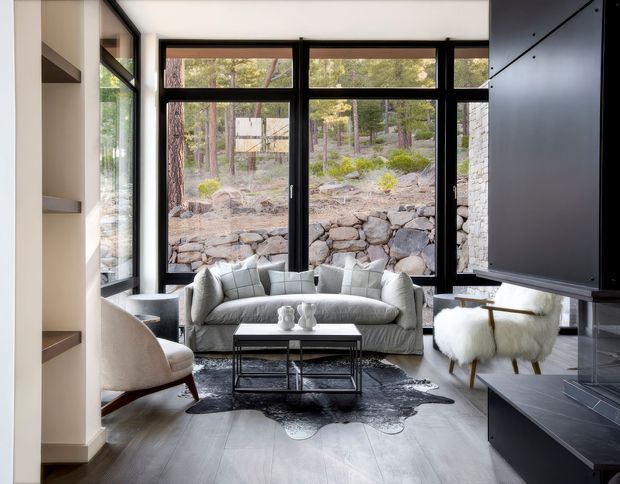 A refined contemporary living room with a grey sofa, sculptural lounge chairs, black metal coffee table, and a dark irregular hide rug. Full-height black-framed glazing opens the room visually to a rocky landscaped setting, while built-in shelving and a large dark fireplace or media surround frame the space. The palette is monochromatic with warm timber flooring and natural stone visible outside.