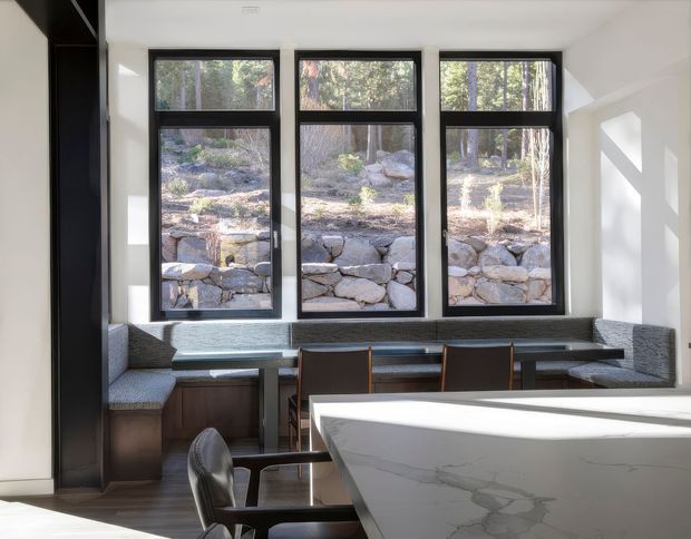 Bright contemporary dining and kitchen nook with three tall black-framed windows overlooking a rocky landscaped slope. A built-in upholstered banquette wraps around a dark timber dining table, while a large white marble-look kitchen island dominates the foreground. The room has white walls, strong natural light, and a clean minimalist composition.