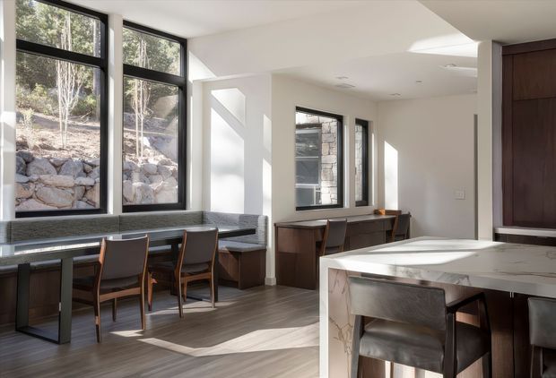 Bright contemporary open-plan kitchen and dining area with extensive black-framed windows, a built-in dining nook, dark timber cabinetry, a marble-look island, and warm timber flooring. The windows overlook a rocky landscaped retaining wall, while strong sunlight creates a calm, refined atmosphere.
