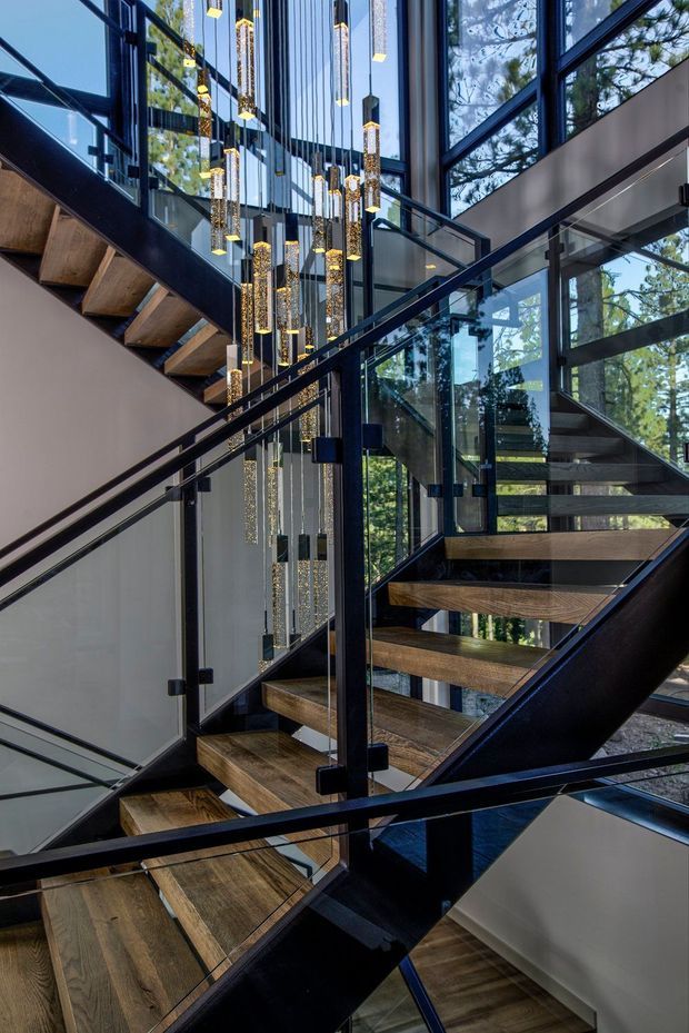 A dramatic multi-level interior stairway with open-riser timber treads, black steel structural framing, and clear glass balustrades. Floor-to-ceiling glazing surrounds the stairwell, reflecting and revealing trees outside. A tall cluster of suspended linear pendant lights forms a striking feature through the stair void. The space is contemporary, sculptural, and filled with natural light.