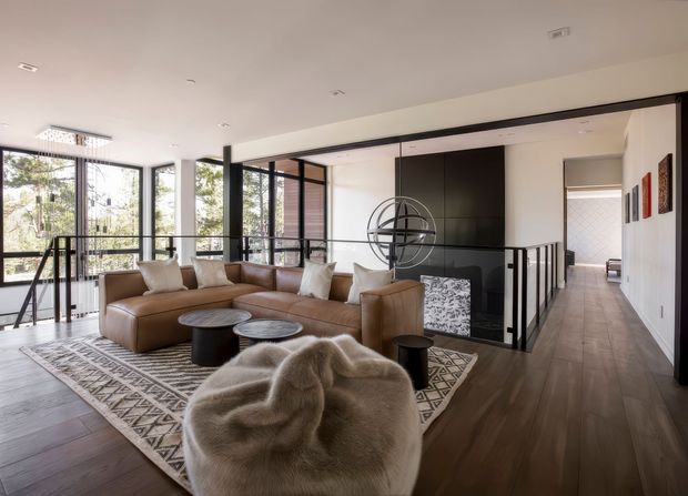 Bright contemporary open-plan living room with floor-to-ceiling black-framed glazing, a large tan sectional sofa, sculptural lounge chair, layered coffee tables, and a geometric area rug. A dark feature wall with an integrated fireplace or media unit separates the living space from a hallway, while black balustrades and timber flooring add architectural definition.